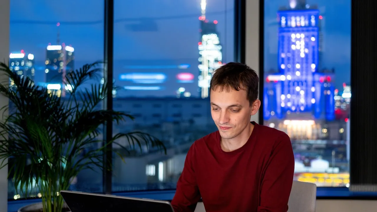 Man in red sweater working on laptop with city skyline lit up at night behind him