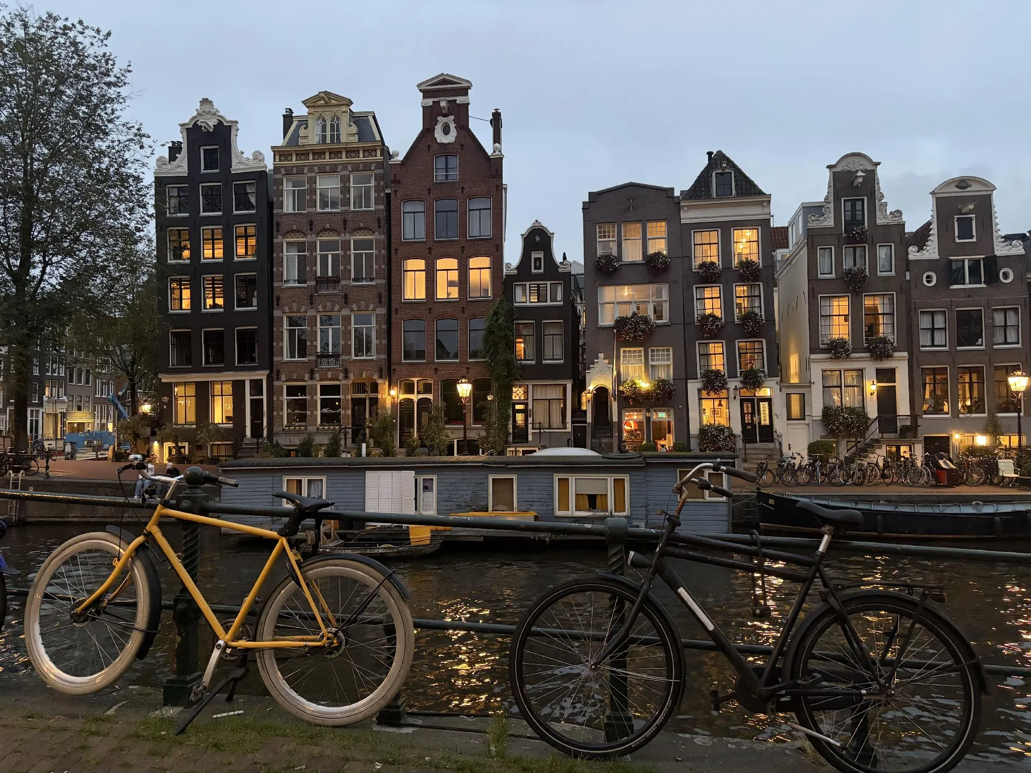 Narrow Amsterdam canal houses with illuminated windows, houseboats on the canal, and bicycles in the foreground.
