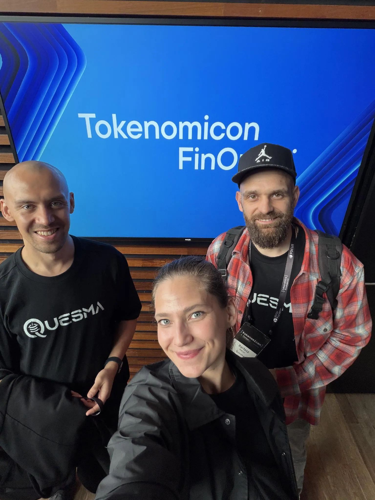 The three Quesma attendees taking a group photo in front of a Tokenomicon and FinOps X display.